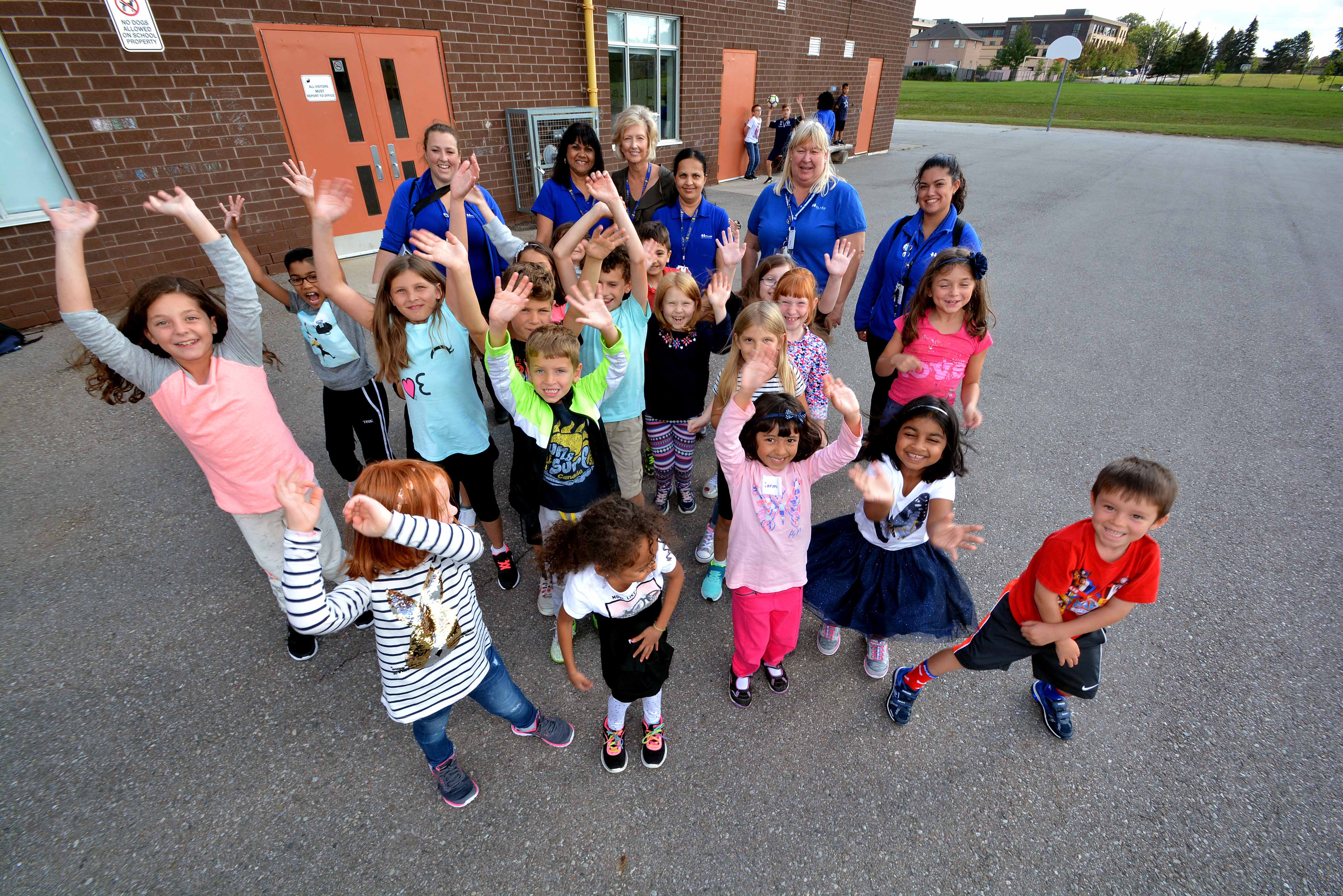 A group of PLASP educators and children stand outside in a school yard with their hands up and big smiles 